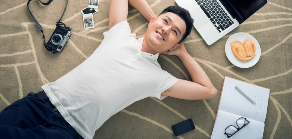 Asian man lies on floor with his laptop and smiles as he thinks